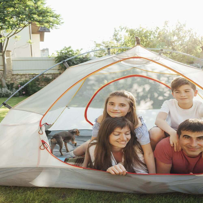 Family enjoying backyard camping in a tent with a barbecue grill in the background on a sunny day