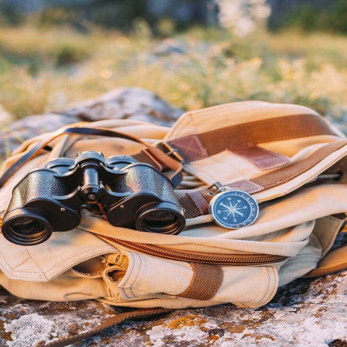 Outdoor adventure backpack with binoculars and a compass on rocky terrain, essential survival gear for wilderness exploration.