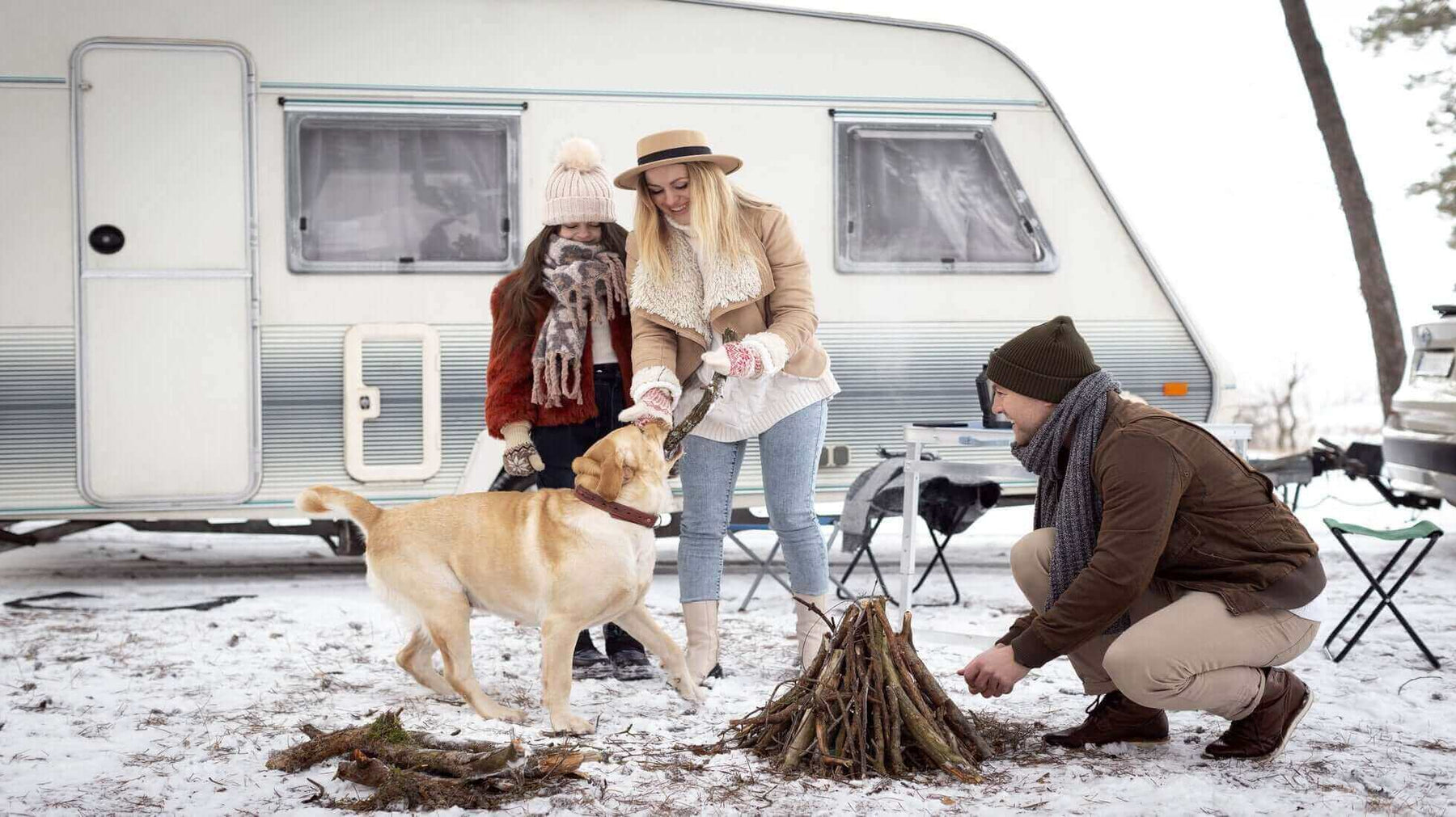 Family camping in the UK, setting up a campfire in the snow beside their caravan with their dog.