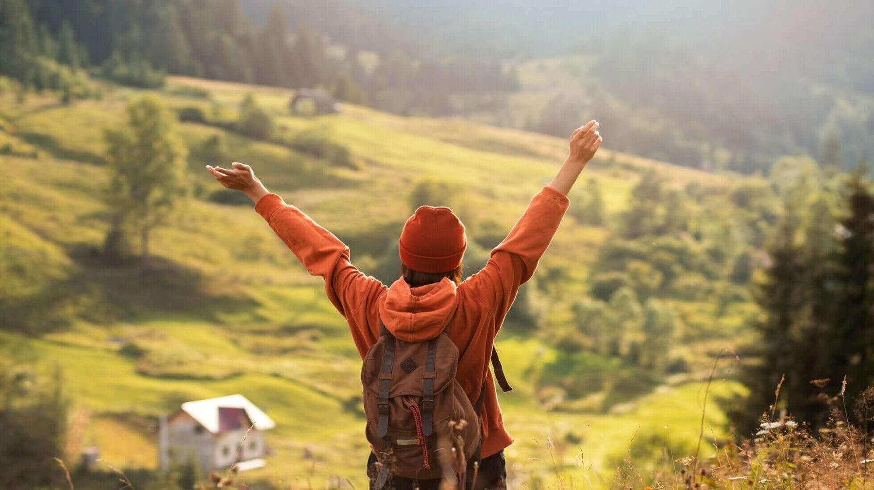 Traveler in red jacket embracing nature on slow travel journey, overlooking scenic green valley and mountains.