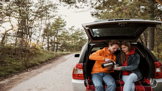 Couple enjoying a hot drink on a road trip, sitting in the back of an open car trunk on a scenic forest road.