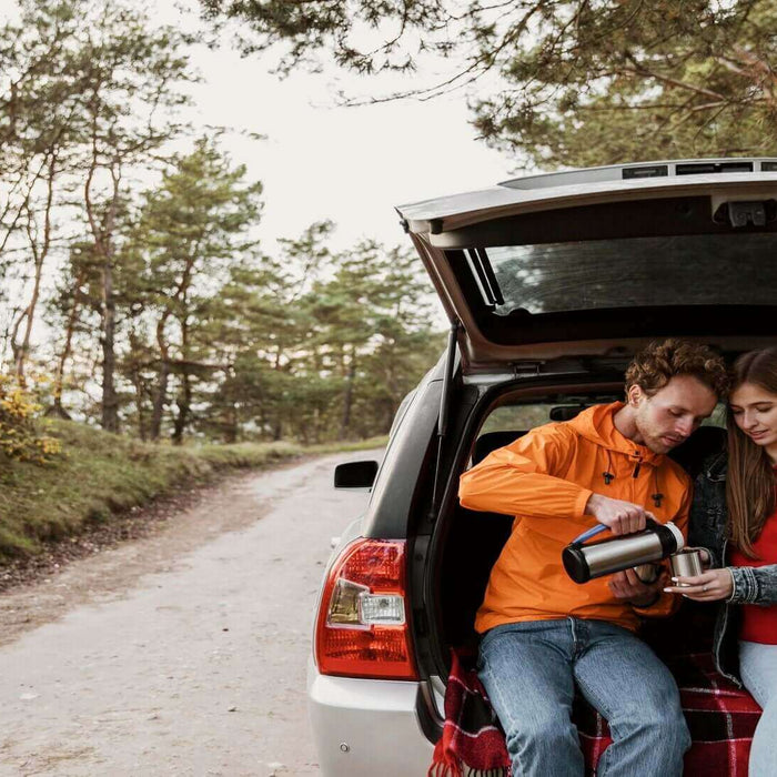 Couple enjoying a hot drink on a road trip, sitting in the back of an open car trunk on a scenic forest road.