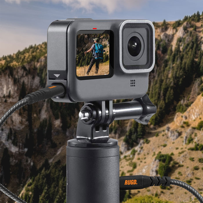 Action camera mounted on a tripod with rugged charging cable, capturing a hiker in a scenic mountain landscape.